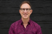 A photo of a white man with glasses wearing a wine-coloured shirt against a dark background