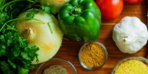 A photo of an opion, green pepper, red tomato, green parsley, white garlic, and three small bowls of orange and yellow spices on a table