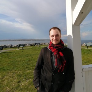 Emmanuel Cuisinier in a jacket and scarf, leaning against a white wood structure, with grass, benches, water, and sky behind him.