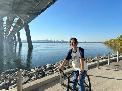 Jiarui Ma on a bicycle next to a body of water with a bridge overhead and the city skyline in the distance.
