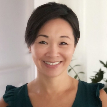 Headshot of woman with black hair smiling in white-painted room with a plant in background