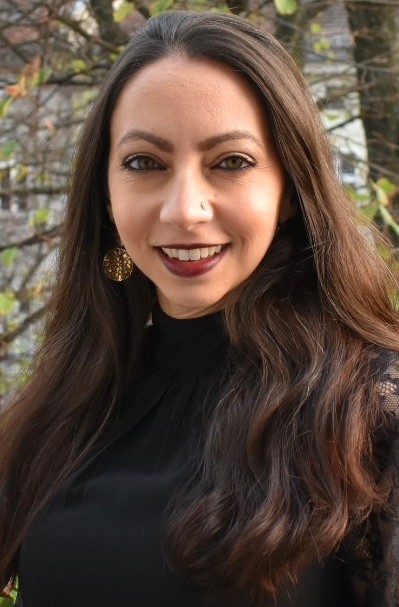 Woman with long brown hair, wearing dark red lipstick, small golden earrings and a black shirt, smiling at the camera. 