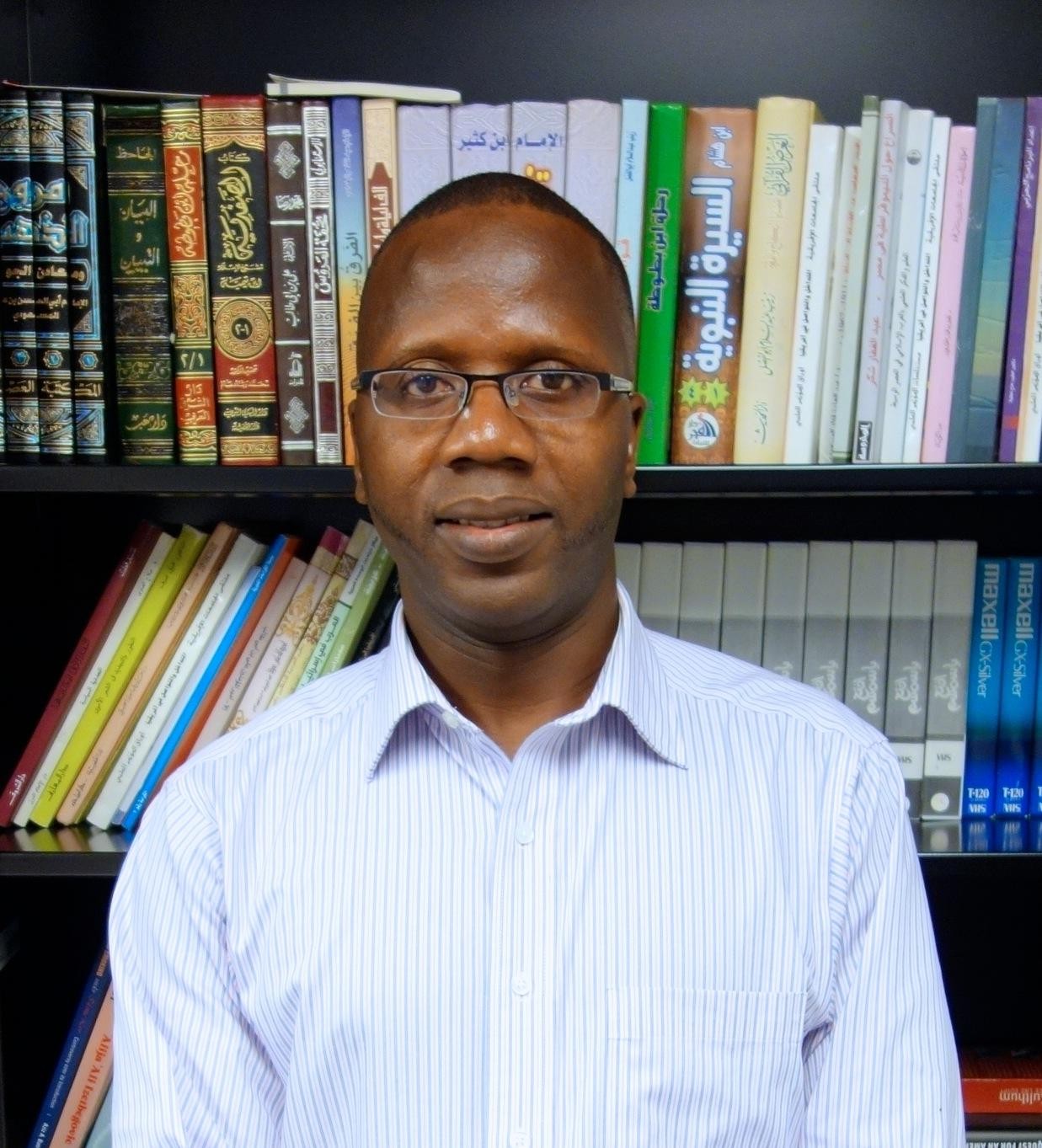 Portrait of Mbaye Lo with bookshelf in background. He wears glasses and a blue button-up shirt.