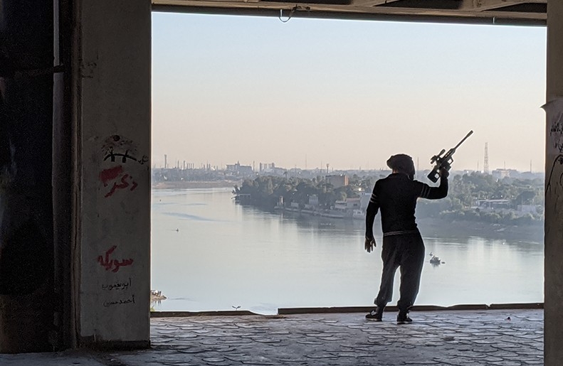 Back of a person holding a paintball gun or prop gun outside a garage with landscape of water and buildings in the background