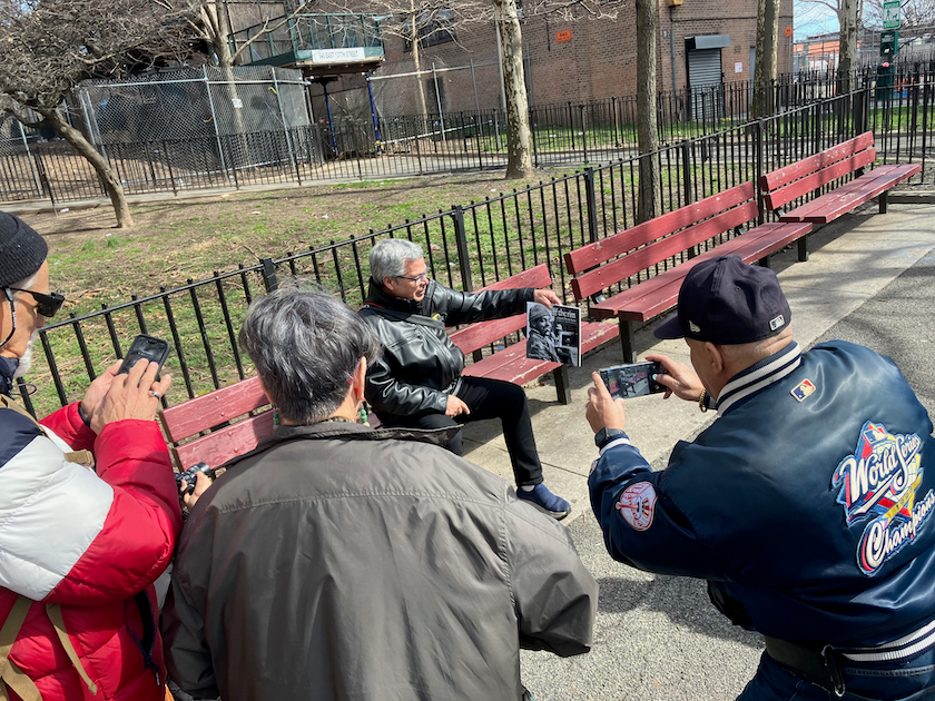 An older man sits on a park bench displaying a book to an audience of three other individuals who take pictures with their iphones.