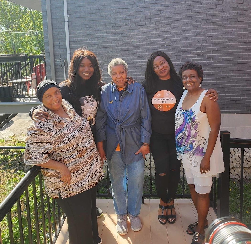 Pictured: Five black women of various ages stand outside on a porch on a sunny day.