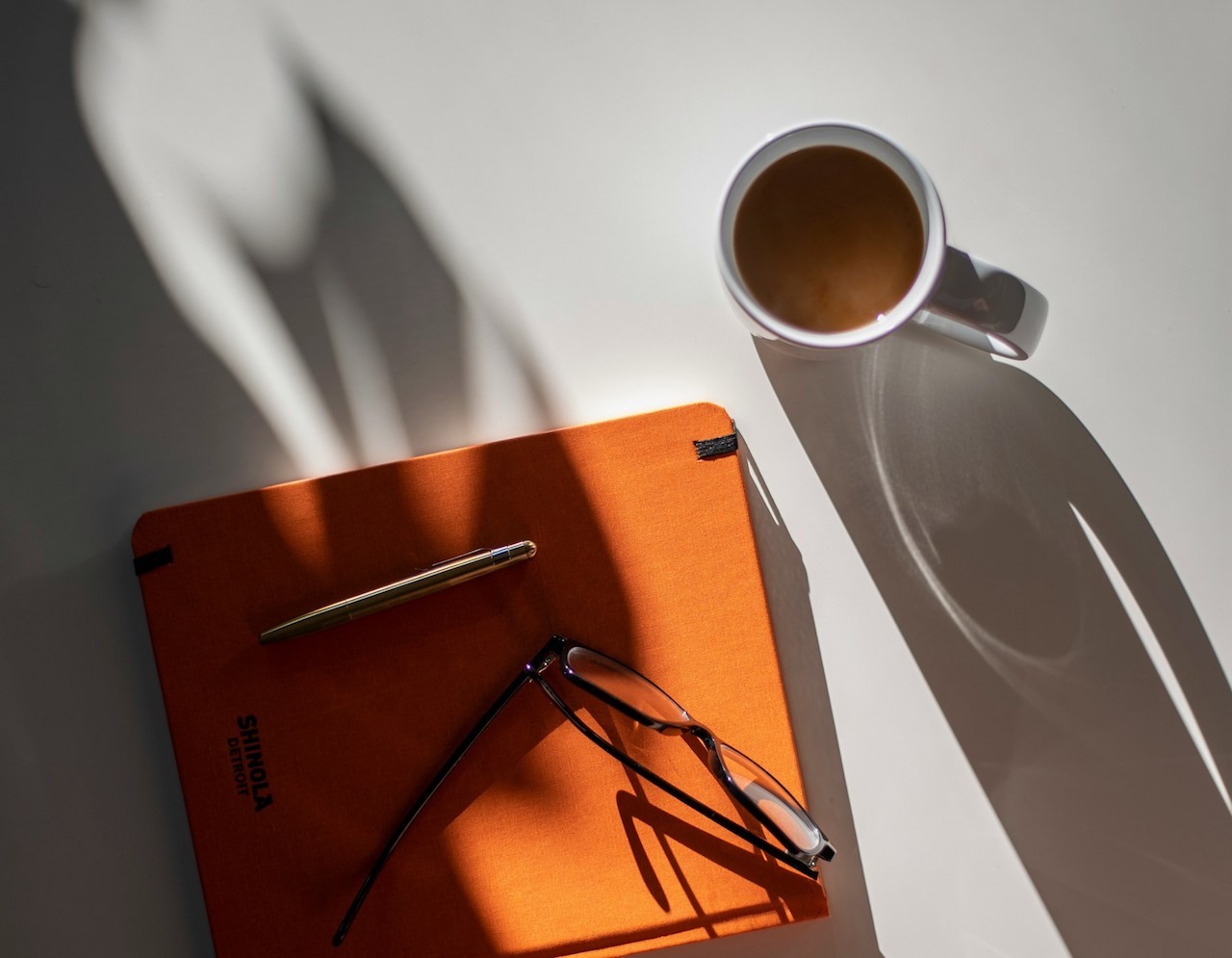 Overhead shot of orange notebook, a pair of glasses which sit on the notebook, and coffee cup.