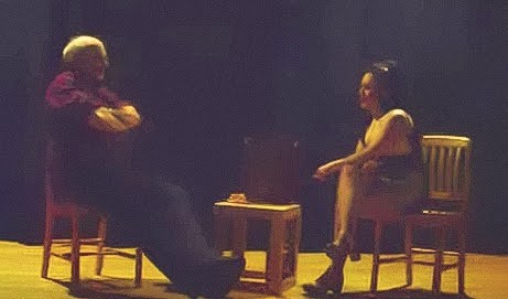 A man and a woman sit facing each other across a small table in a dimly lit room. 