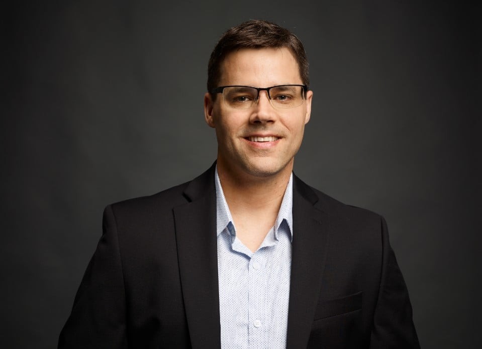 Cory Labrecque stands in front of dark gray background. He wears a black suit coat over a blue button-up shirt and wears black square-framed glasses. He has short brown hair.