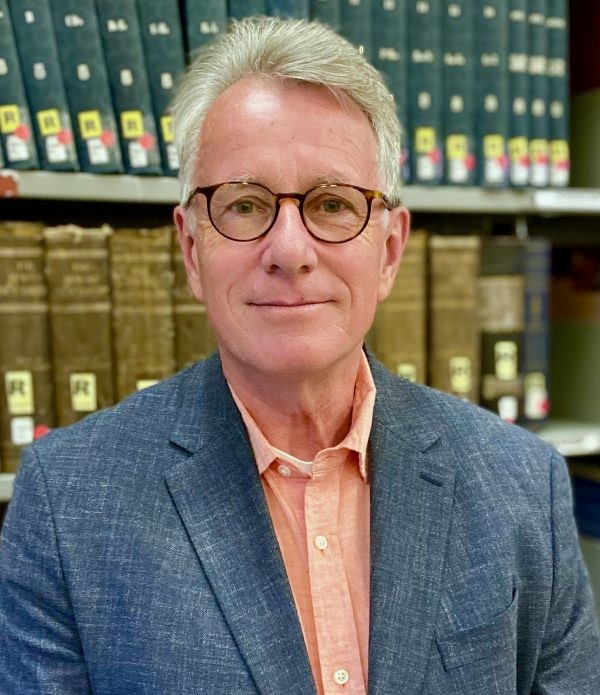 Gerbern S. Oegema poses in front of a shelf of books. He wears a blue suit coat over a salmon pink button-up shirt. He wears oval tortoise-shell glasses and has white hair.