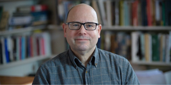 Charles Briggs in front of shelves of books. His head is shaved and he wears rectangular black framed glasses. He wears a navy blue button-up shirt.