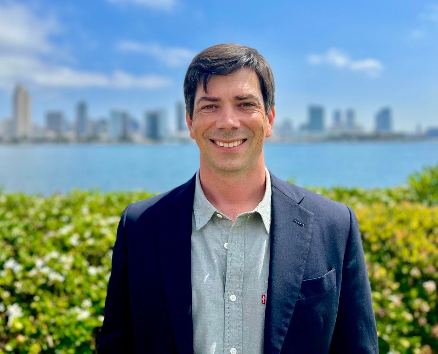 Eric Morelli with green bushes, body of water and distant cityscape in the backgroud. He wears a blue suit coat over a light blue button-up shirt. He has short brown hair parted on the left side.