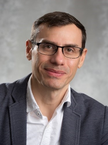 Headshot of man with black glasses and brown hair, wearing a grey blazer and white button-up shirt.