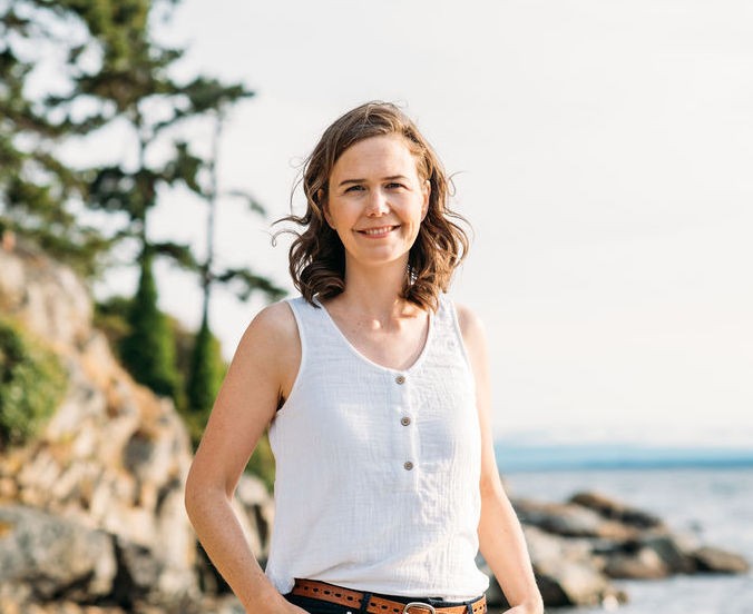 Deborah stands against a backdrop of a shoreline with trees and water. 