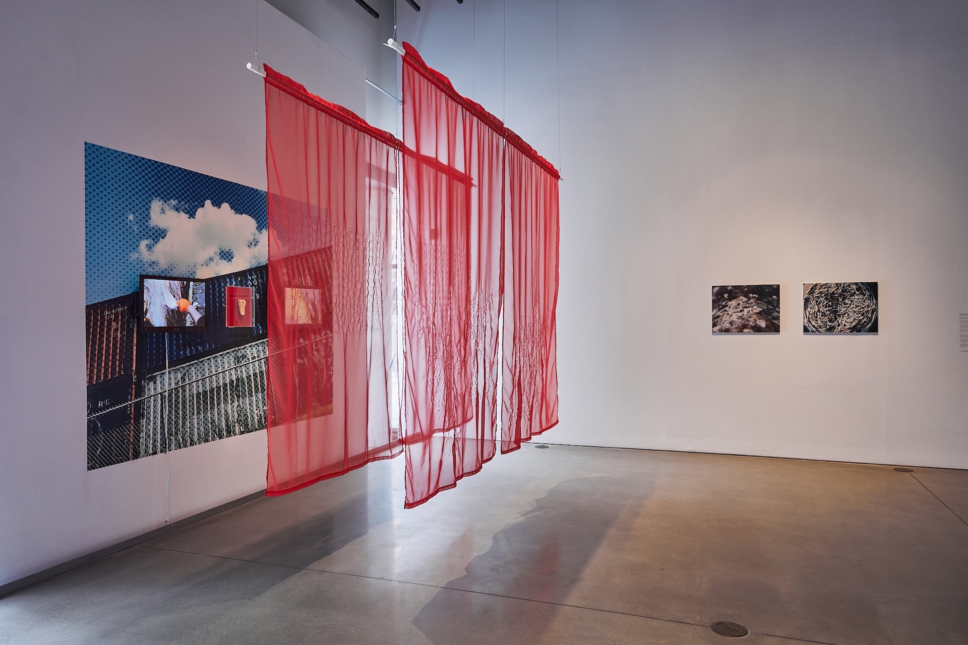 photograph of a gallery with two white walls visible. The wall in front has two printed images. The wall to the left has a large-scale print of an urban landscape, overlayed by two screens. In front of that wall is two layers of sheer red fabric, acting as curtains protecting the work.