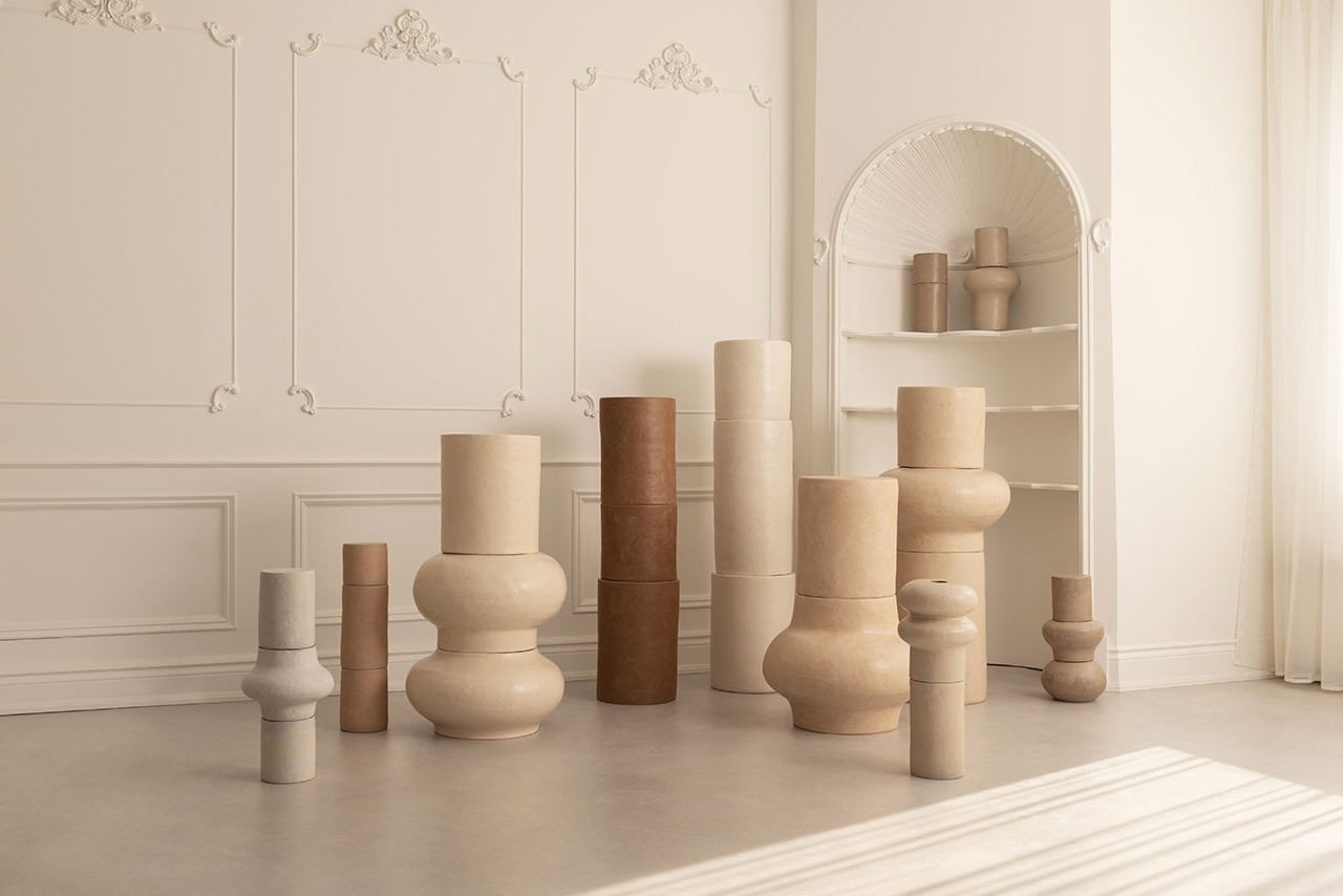 A minimalist room displays various neutral-toned ceramic sculptures of different shapes and sizes against an ornate white wall and built-in shelf.