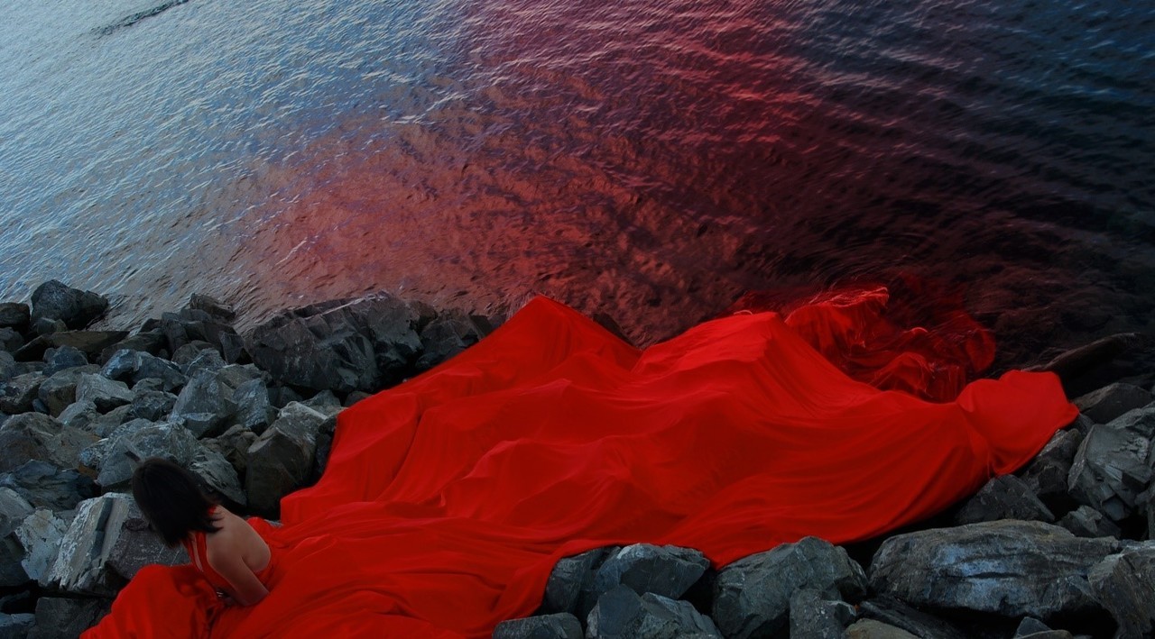 Woman sitting on rocks near water, wearing bright red dress with long trail 