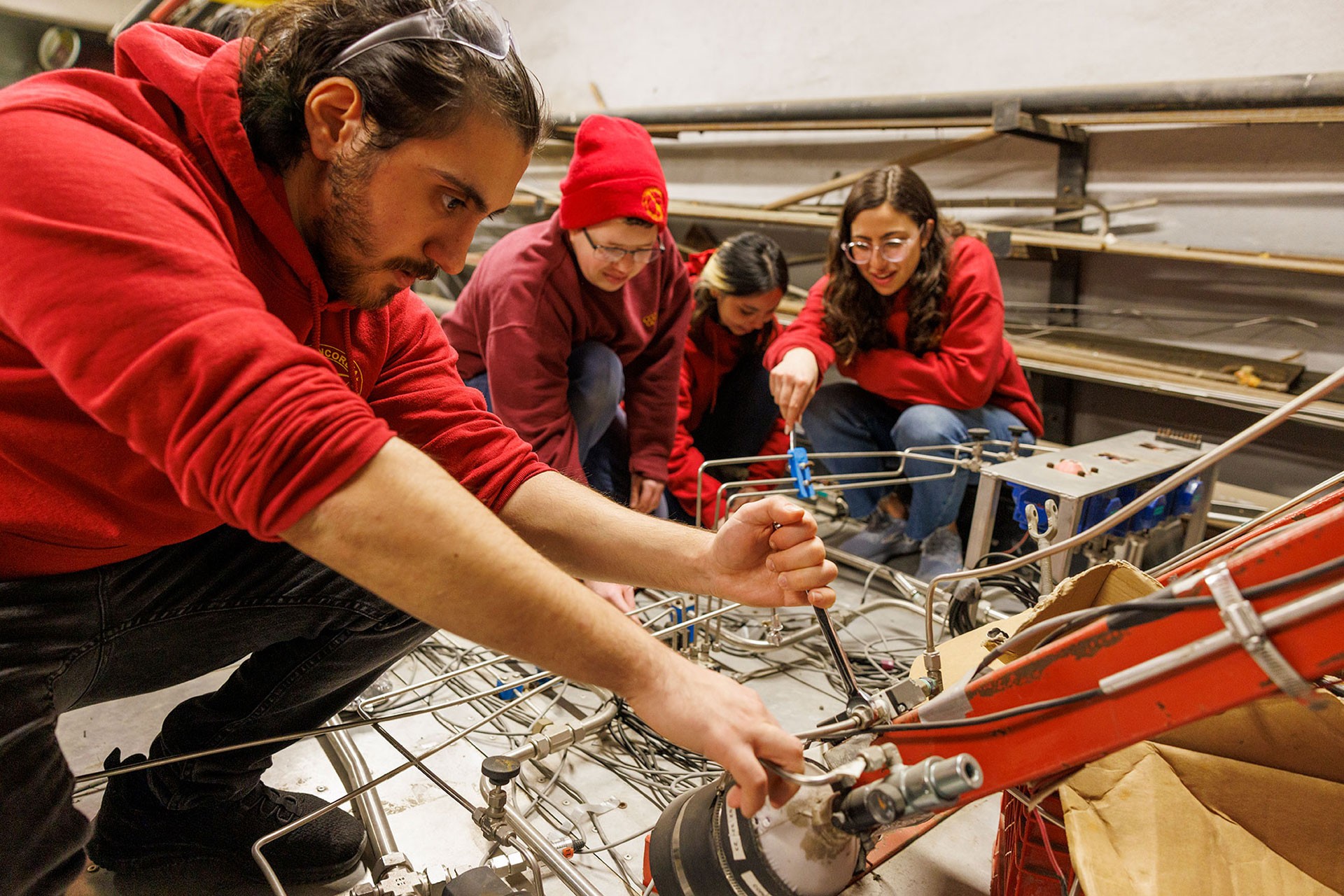 Four Concordia students in red hoodies and safety glasses work closely on a rocket propulsion system in a lab, adjusting valves and wiring on the ground.