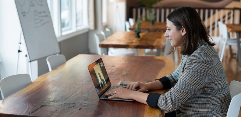 Woman wearing blazer on virtual phone call, seated in a cafe or working space with wooden tables and white chairs