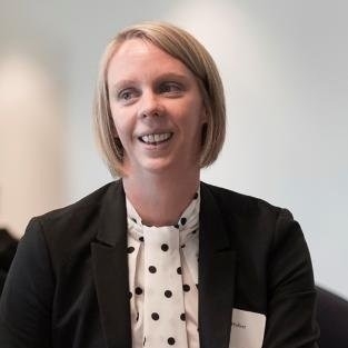 Woman with short light hair smiling, wearing white and black polka-dotted blouse and black blazer