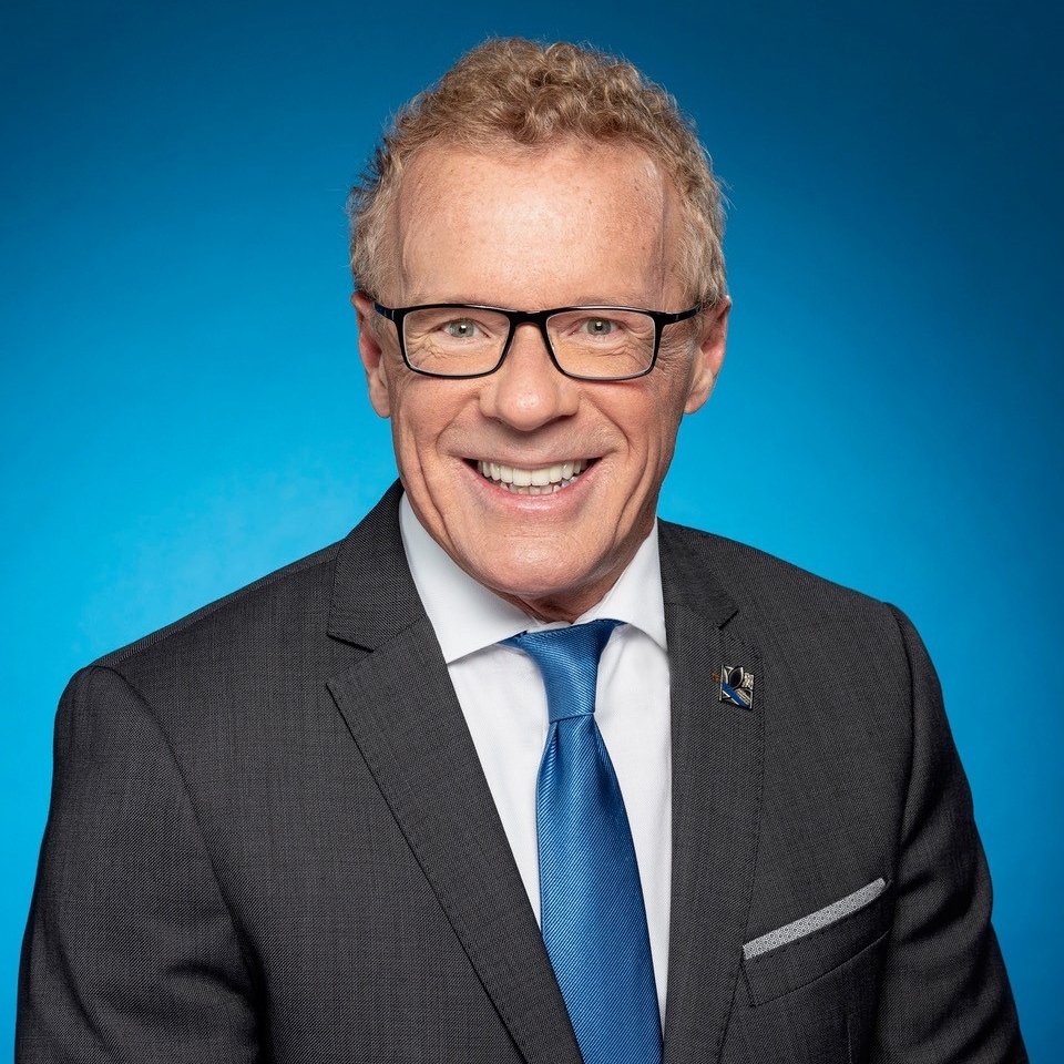Headshot of smiling man with glasses wearing suit and blue tie in front of a vibrant blue background