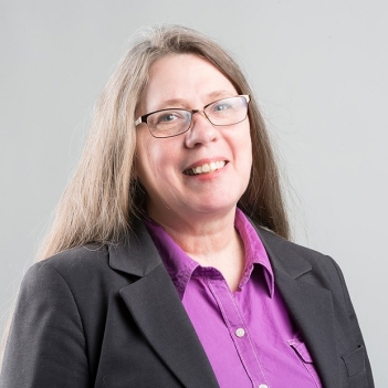 Janis Timm-Bottos photographed against gray background. She has long grayish-brown hair and is wearing glasses, a purple button-down and a black blazer.