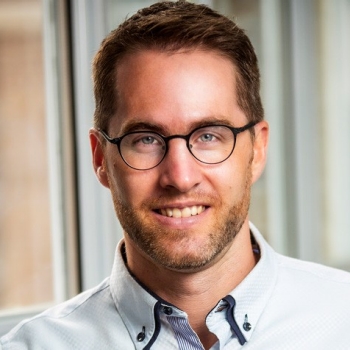 Jean-Philippe Gouin is looking straight at the camera and smiling. He is wearing black, oval-shaped glasses and a light blue button-down shirt.