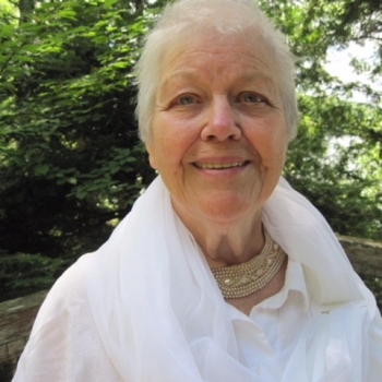Marguerite Dorion is pictured outdoors with green leafy trees in the background. She has short cropped white hair and is wearing a white button-down shirt and a white scarf.