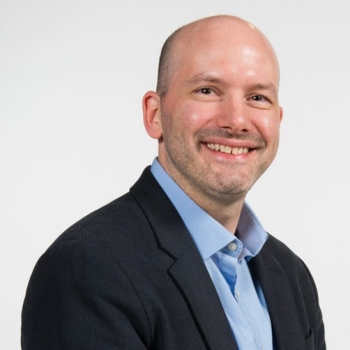 Simon Bacon is photographed against a gray background. He is smiling at the camera and wearing a blue button-down shirt with a black blazer.