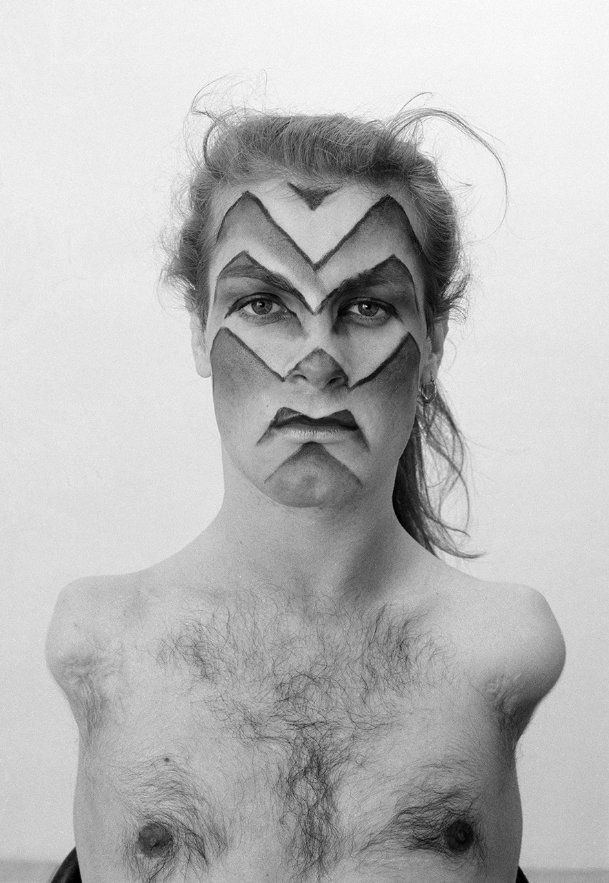 Black-and-white image of a person from the chest up with face paint and no shirt on.