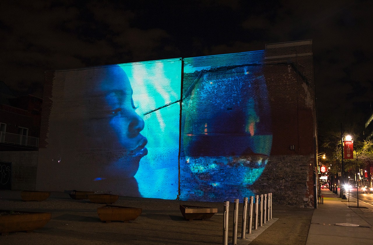 The side of a building lit with the projection of the face of a young Black woman.
