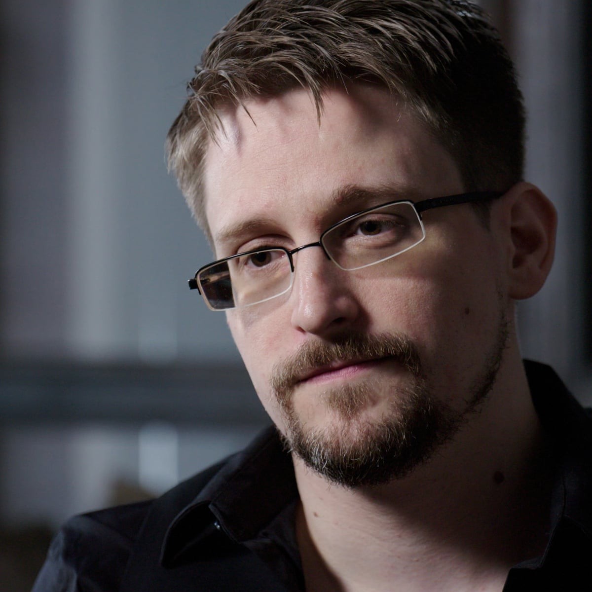 Headshot of Edward Snowden from shoulders up. White man with short-cropped brown hair, wireframe glasses and goatee. Wearing black, button-up shirt.