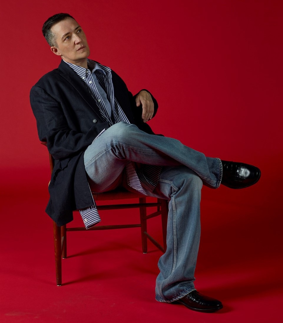 Person wearing navy blazer and blue-striped collared shirt and light-blue jeans sitting in wood chair in front of muted red backdrop. 