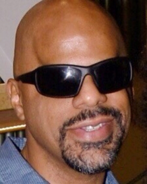 Thomas Reid a brown skin Black man with a clean shaven bald head smiles into the camera. He has a goatee and is wearing dark shades and a gray button up shirt.