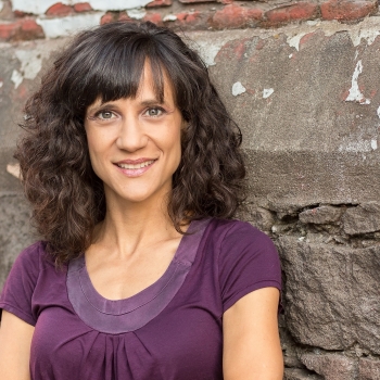 A white woman with olive complexion and curly dark brown hair smiles for the camera. She wears an eggplant purple shirt and leans against a rough stone wall.