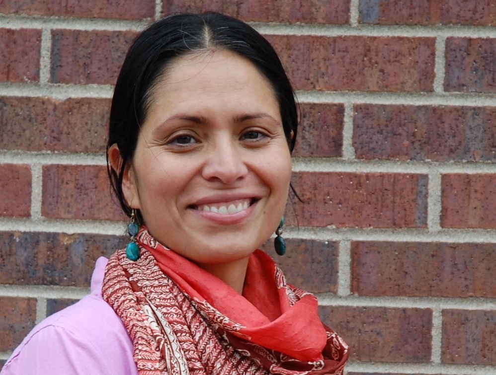 Smiling dark-haired woman wearing colourful patterned orange-and-white scarf and light-purple shirt in front of brick wall