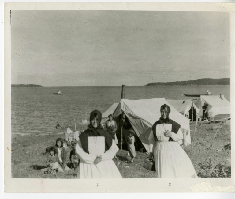 Black-and-white image of two nuns and children on open landscape near water