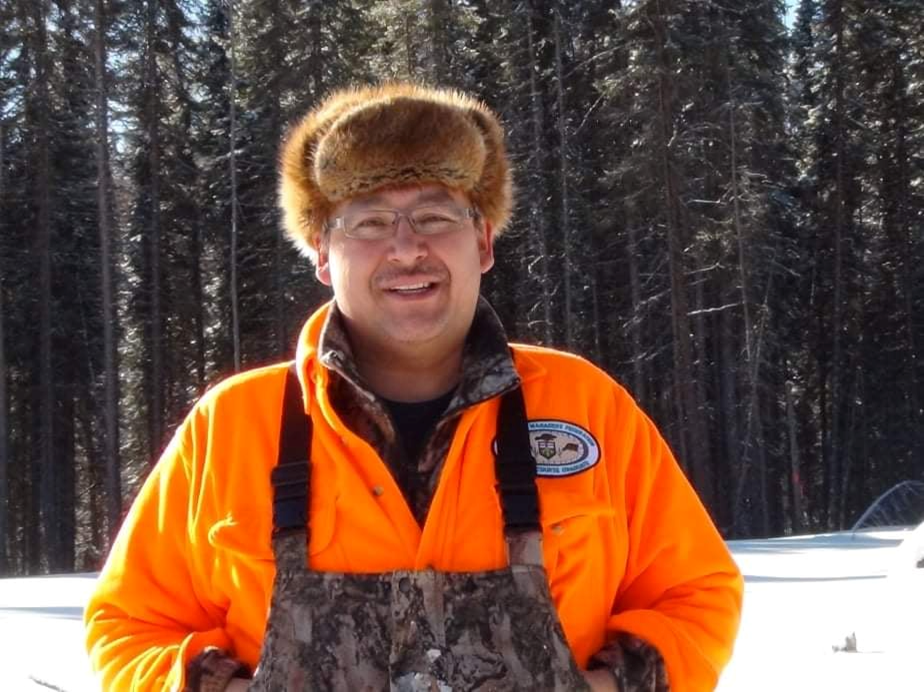 Richard Wesley, Traditional Ecological Knowledge Keeper, Constance Lake Anishinaabe. He is wearing a fur cap, glasses, orange jacket and black overalls and stands in the winter woods