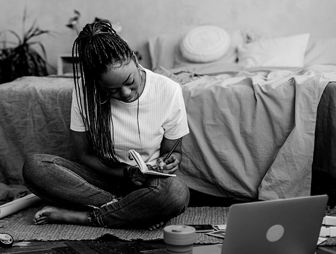 Black woman seated on bedroom floor writing in a notebook