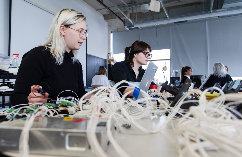 Two women sit at a desk with a laptop and many wires in the foreground.