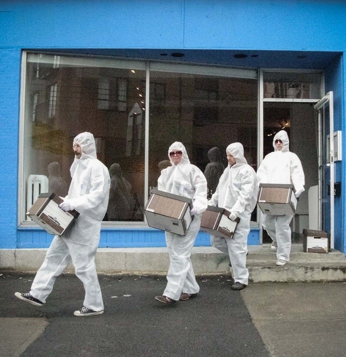 4 people wearing white hazmat-looking suits and sunglasses, carrying banker's boxes out of building