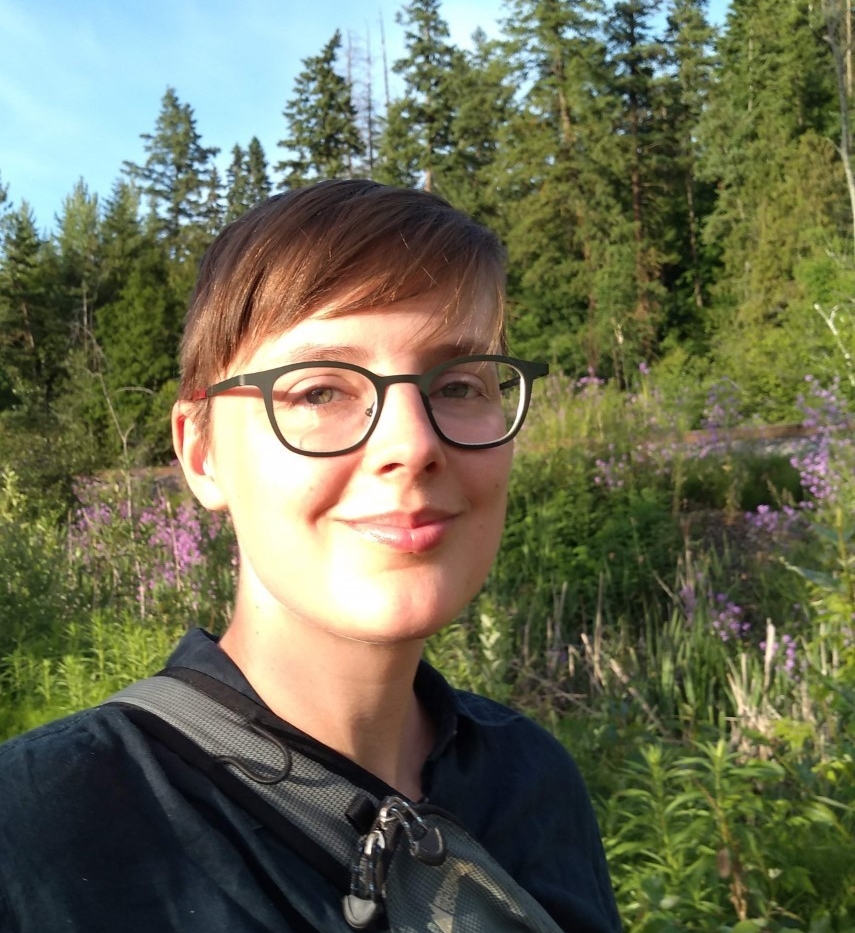 A young, smiling woman with short hair and glasses standing in a clearing surrounded by trees on a sunny day