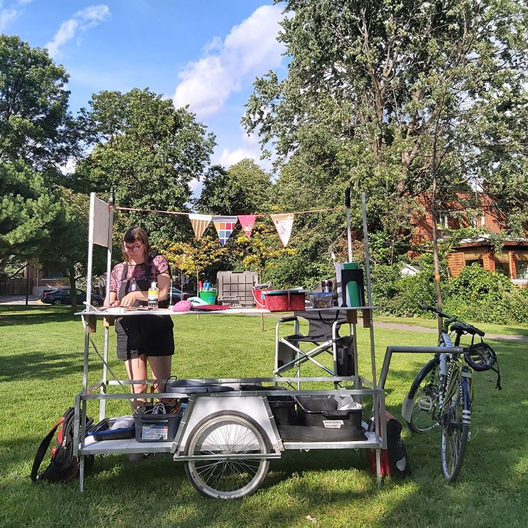 A large bike trailer that facilitates art making. On a sunny day on the grass in a park in NDG.