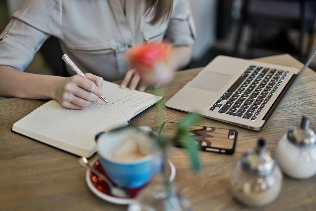 Close up of a woman's hand writing in a notebook, next to a laptop, phone and cup of coffee