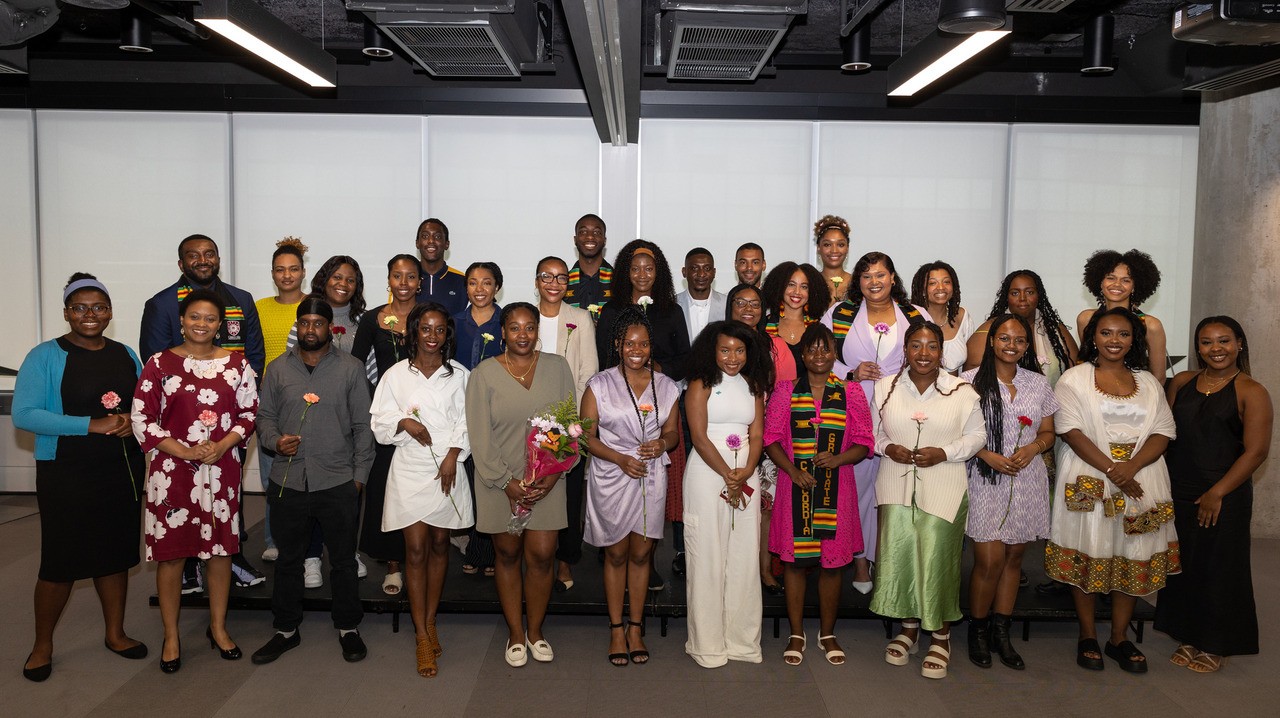 Standing group shot of women and men in formal attire, most of whom are each holding a flower.