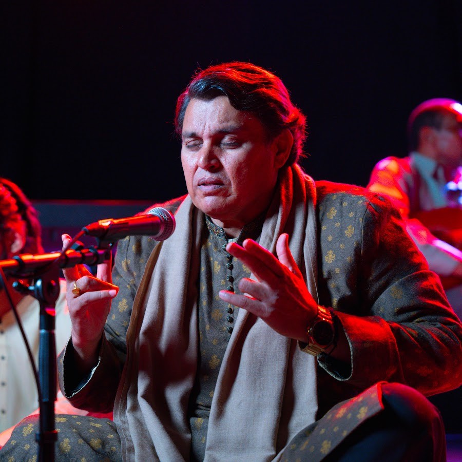 A man in traditional attire performs on stage with closed eyes, expressing emotion while singing into a microphone. The scene is bathed in warm red and blue lighting.