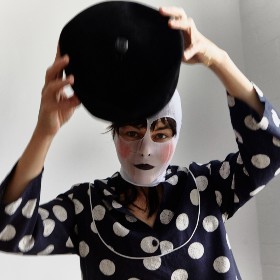 A woman in a mask wears a polka dot shirt and holds an object over her head