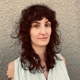 A woman with medium-length, dark hair stands in front of a wall