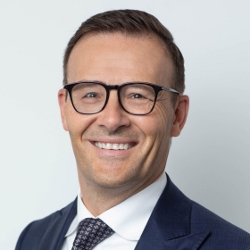 A man with short brown hair and glasses wears a checkered tie and smiles for the camera