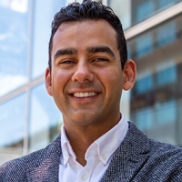 A man with short black hair wears a tweed jacket and white shirt